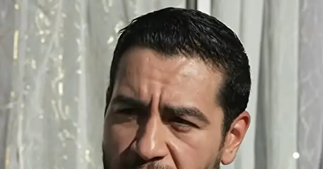 The image shows a close-up of a person with a serious expression, set against a backdrop of light-colored curtains.
