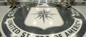 The image shows the logo of the Central Intelligence Agency (CIA) on the floor, with individuals walking past it.