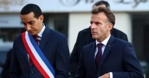 The image shows two men in formal attire, one wearing a red, white, and blue sash, walking together in what appears to be a public or official setting.