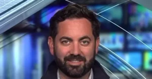 The image shows a man with a beard and short hair, wearing a dark jacket, in what appears to be a news studio setting.