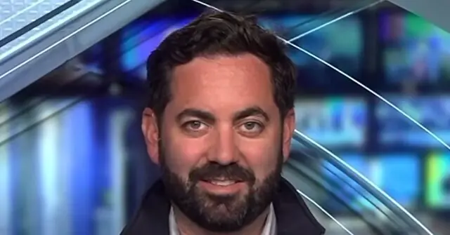 The image shows a man with a beard and short hair, wearing a dark jacket, in what appears to be a news studio setting.
