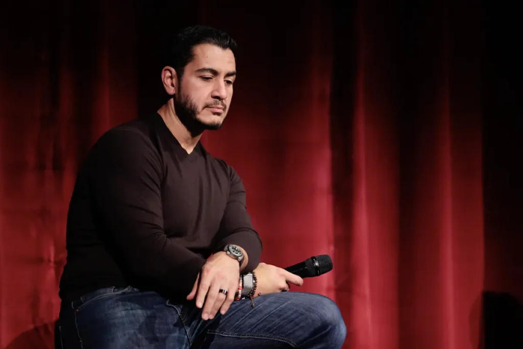 A man is seated on stage, holding a microphone, with a red curtain in the background.