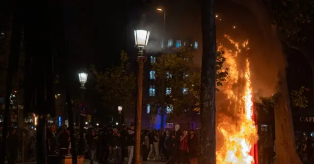The photo depicts a nighttime scene with a crowd gathered around a large fire, illuminated by streetlights and the glow of the flames.