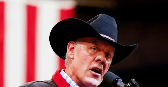 The image shows a man wearing a cowboy hat and a red shirt, speaking into a microphone in front of an American flag.