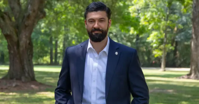 A man in a suit stands outdoors in a park-like setting with trees in the background.