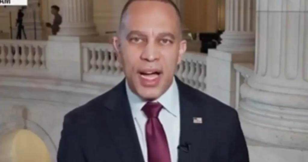 The image shows a man in a suit and tie speaking in front of a backdrop featuring classical architecture, likely a government building.