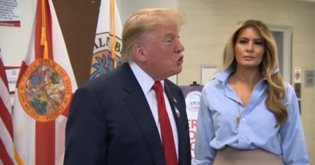 The image shows a man in a suit and a woman in a light blue blouse standing in front of flags, possibly at a public event or press conference.
