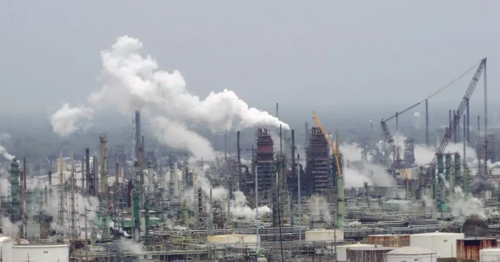 The image shows a large industrial facility with numerous smokestacks emitting plumes of smoke, indicating ongoing industrial activity and potential environmental impact.