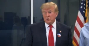 The image shows a man in a suit and red tie standing in front of an American flag, likely during a formal or official event.