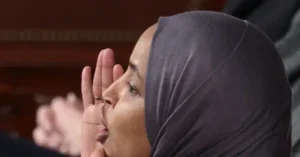 A woman wearing a hijab is shown in a close-up, with her hands raised in a gesture of prayer or reflection.