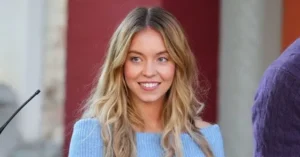The image shows a person with long, wavy blonde hair wearing a light blue sweater, standing in front of a microphone and a red and white background.
