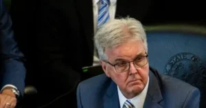 The image shows a man in a suit and tie, seated at a desk with a microphone, appearing to be in a formal setting, possibly a press conference or official meeting.