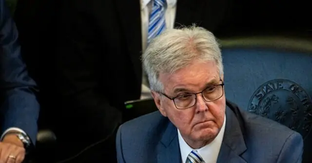The image shows a man in a suit and tie, seated at a desk with a microphone, appearing to be in a formal setting, possibly a press conference or official meeting.