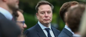 The image shows a man in a suit and tie, surrounded by other individuals, in what appears to be a formal or professional setting.
