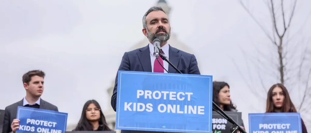 A man speaks at a podium with a sign that reads "PROTECT KIDS ONLINE," surrounded by individuals holding similar signs, in front of a backdrop featuring the U.S. Capitol building.