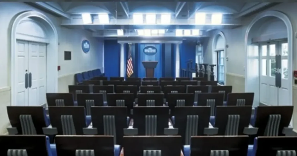 The image shows an empty press briefing room with rows of chairs facing a podium and a U.S. flag.