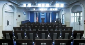 The image shows an empty press briefing room with rows of chairs facing a podium and a U.S. flag.