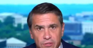 The image shows a news anchor in a studio setting with a blurred background of the White House.
