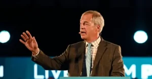 A man in a suit and tie is speaking at a podium with a microphone, gesturing with his right hand.