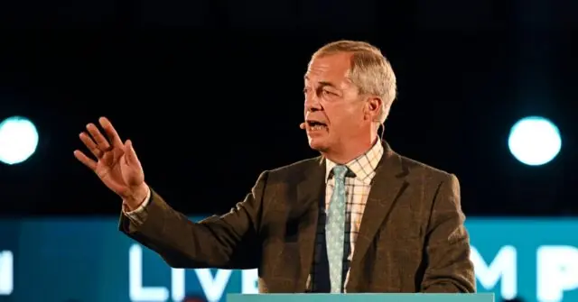 A man in a suit and tie is speaking at a podium with a microphone, gesturing with his right hand.