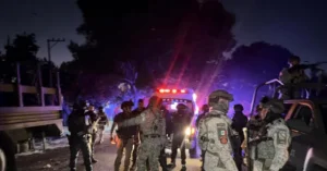 The image depicts a nighttime scene with law enforcement and military personnel gathered around a vehicle, illuminated by emergency lights and red laser beams, suggesting a tense or active situation.