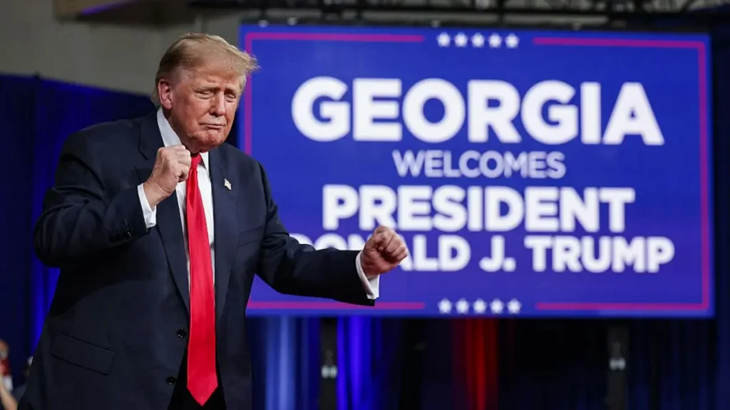 The image shows President Donald J. Trump at a campaign event in Georgia, with a sign welcoming him.