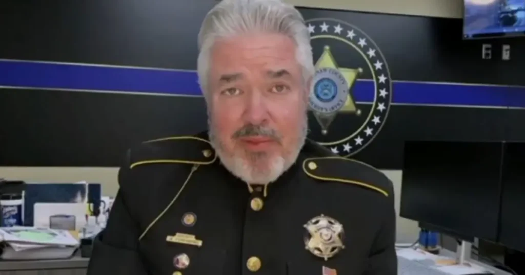 The image shows a person in a police uniform standing in front of a backdrop featuring a police department emblem and a blue stripe.
