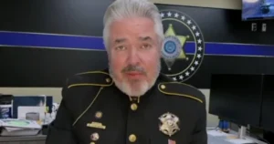 The image shows a person in a police uniform standing in front of a backdrop featuring a police department emblem and a blue stripe.