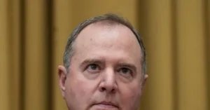 The image shows a close-up of a man with a serious expression, set against a blurred background.
