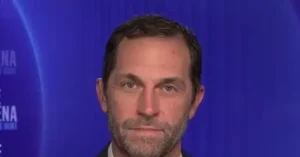 The image shows a man with a beard and short hair, wearing a suit, against a blue background with text partially visible.