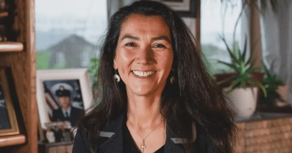 The image shows a smiling woman with long dark hair, wearing a black top and a cross necklace, in a warmly lit indoor setting with framed pictures and plants in the background.