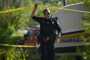 A police officer stands at a crime scene, holding a yellow caution tape, with a forensic van in the background.