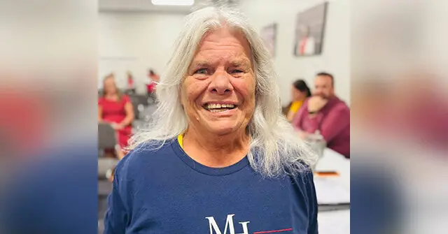 The image shows a smiling individual with long white hair, wearing a blue shirt, in what appears to be a casual indoor setting with other people in the background.