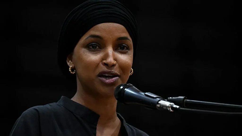 A woman wearing a black turban and earrings speaks into a microphone against a dark background.