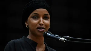 A woman wearing a black turban and earrings speaks into a microphone against a dark background.