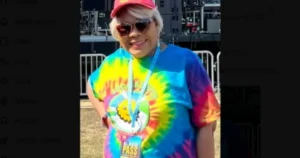 A person wearing a colorful tie-dye shirt and a red cap is standing outdoors, smiling, with a lanyard around their neck.
