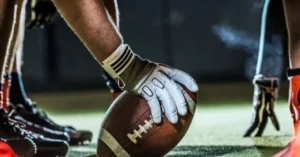 A football player prepares to hold the ball during a game, wearing gloves and cleats.