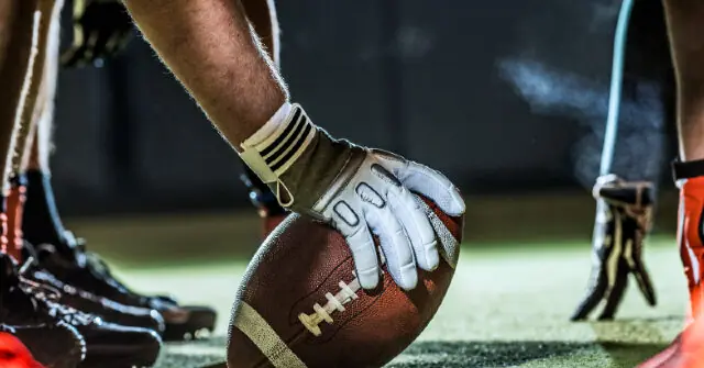 A football player prepares to hold the ball during a game, wearing gloves and cleats.