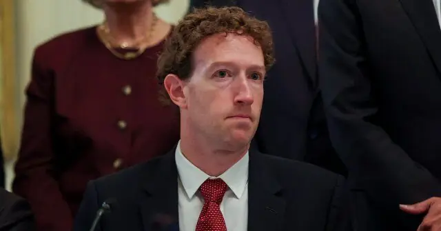 The image shows a man in a suit and tie, seated at a table with a microphone, appearing to be at a formal event or hearing.