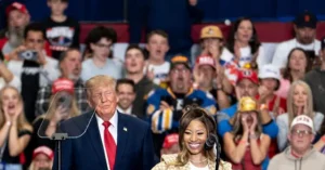 The image shows a public event with a crowd of people in the background, some wearing red hats, and a man in a suit standing at a podium with a woman beside him.