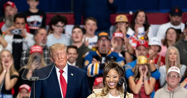 The image shows a public event with a crowd of people in the background, some wearing red hats, and a man in a suit standing at a podium with a woman beside him.