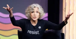 The image shows a person on stage wearing a black shirt with the word "resist" on it, gesturing with their hands outstretched.
