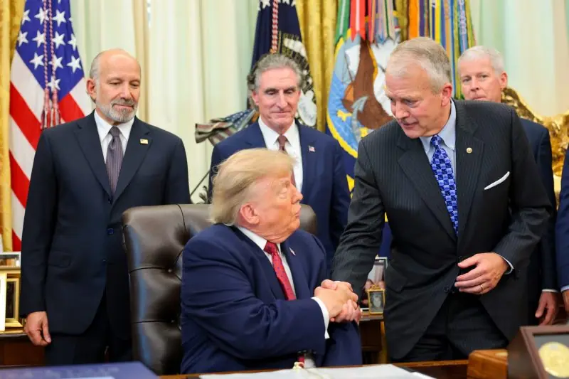 The image shows a formal meeting in the Oval Office with President Trump seated at his desk, shaking hands with another man, while other officials stand in the background.