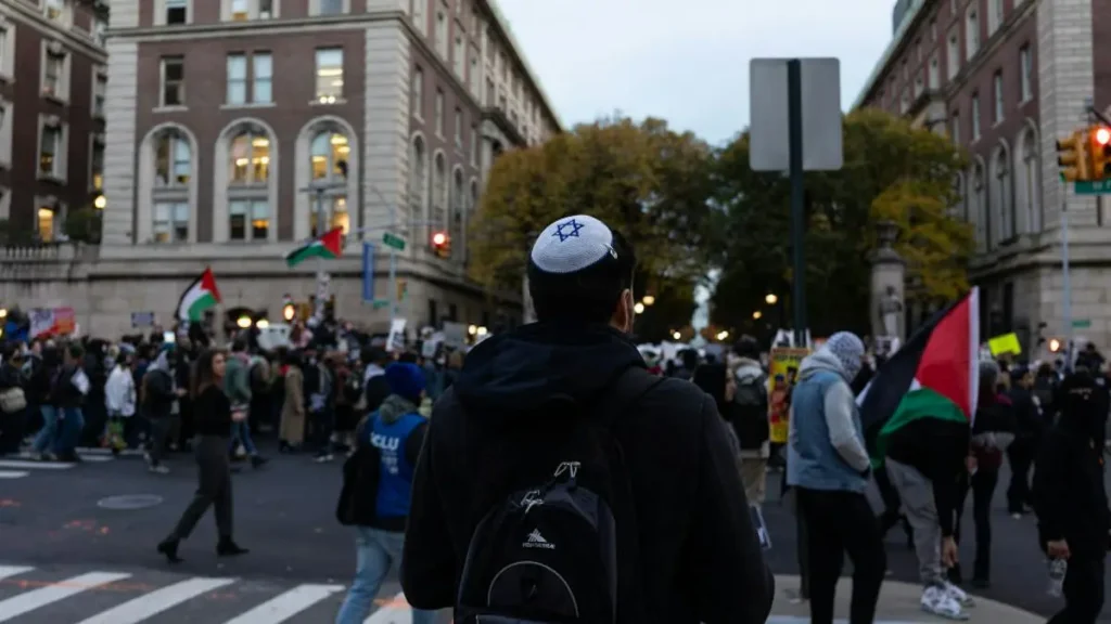 A person wearing a kippah and backpack walks through a crowded street scene with protesters holding Palestinian flags.