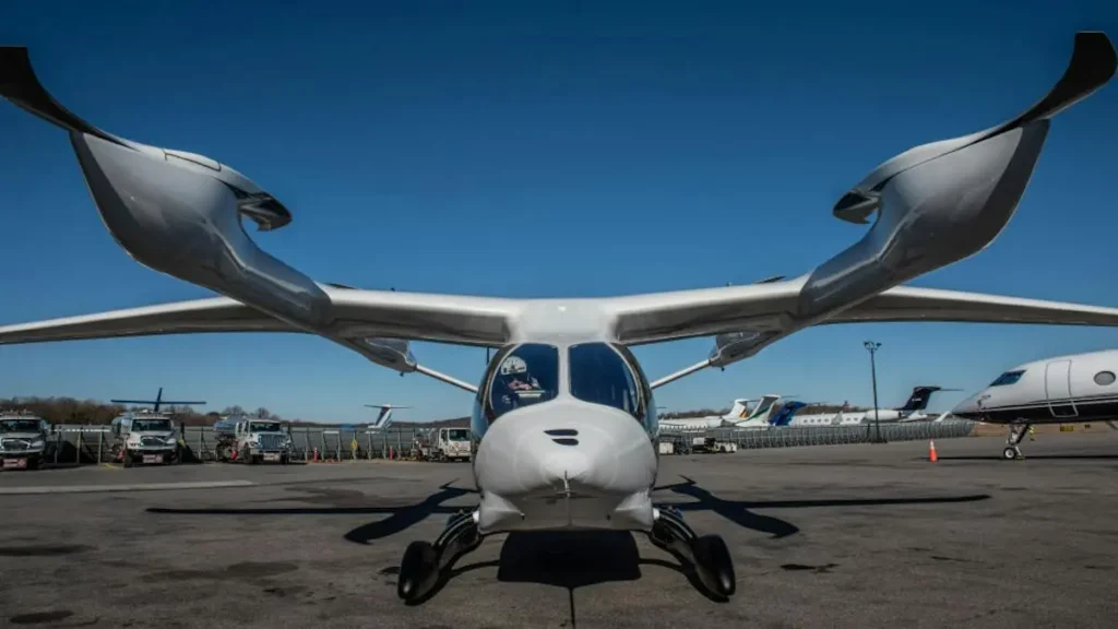 The image shows a futuristic-looking aircraft with a unique design, featuring large, swept-back wings and a sleek, modern body, parked on an airport tarmac.