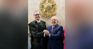 The photo shows two individuals shaking hands in front of a large, ornate emblem, suggesting a formal or official setting.