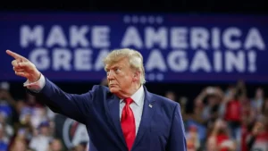 The image shows a person in a suit and red tie pointing forward with a "MAKE AMERICA GREAT AGAIN!" sign in the background.