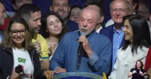 A man speaks into a microphone surrounded by a group of people, with a Brazilian flag and the phrase "Ordem e Progresso" visible in the foreground.