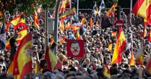 The photo depicts a large crowd gathered outdoors, waving Spanish flags and holding a sign with a caricature of a person, suggesting a political rally or demonstration.