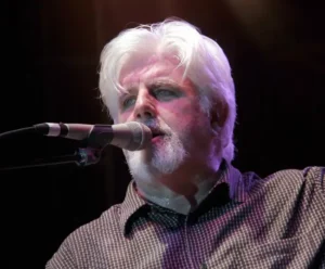 The image shows a person with white hair and a beard, wearing a checkered shirt and a bow tie, singing into a microphone on stage.